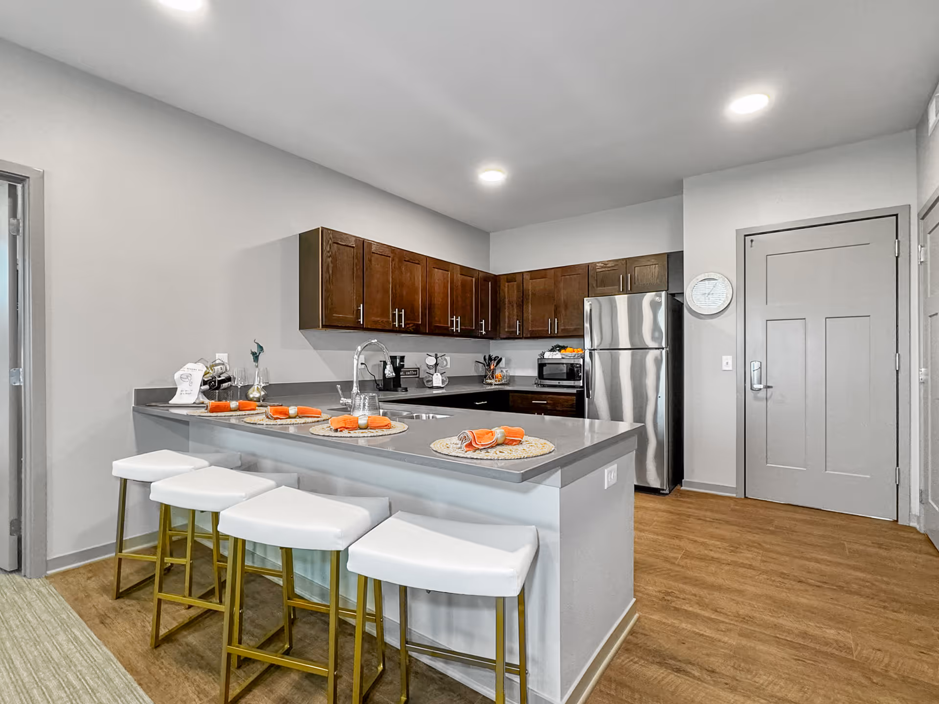 Modern kitchen with dark wood cabinets, stainless steel refrigerator, and a gray countertop island with four white bar stools. The island is set with placemats and orange napkins. The floor is wood, and the walls are painted light gray. There is a door and a clock on the wall near the refrigerator.