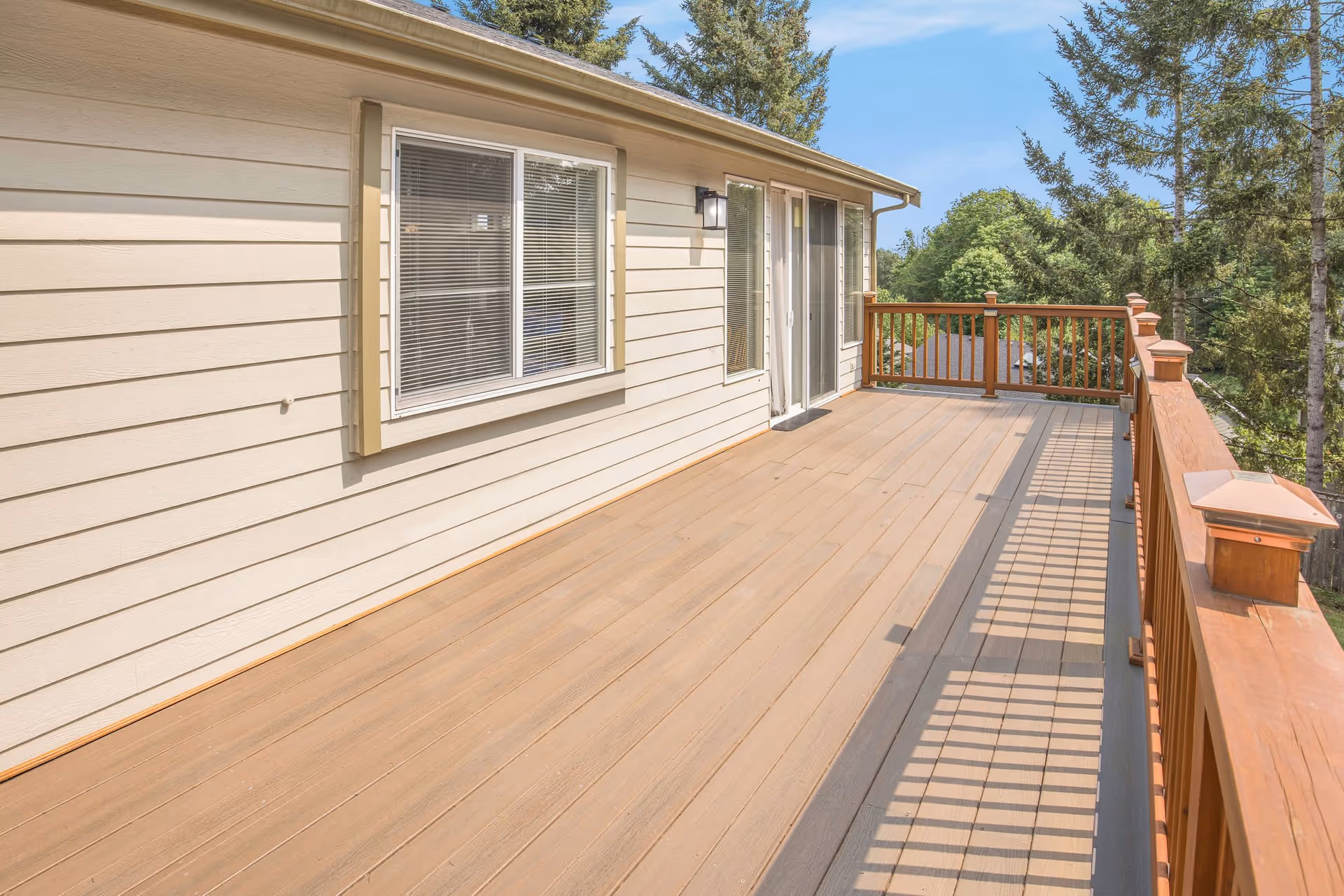 A spacious wooden deck attached to a beige house with horizontal siding. The deck has a wooden railing with vertical balusters and decorative post caps. There are two windows with blinds and a sliding glass door leading inside. Trees and greenery are visible in the background under a clear blue sky.