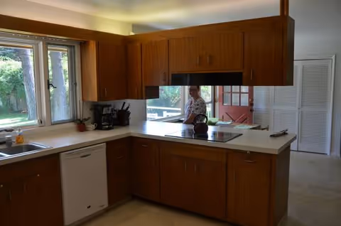 Wooden kitchen with countertops and appliances; a person stands near a pass-through window.