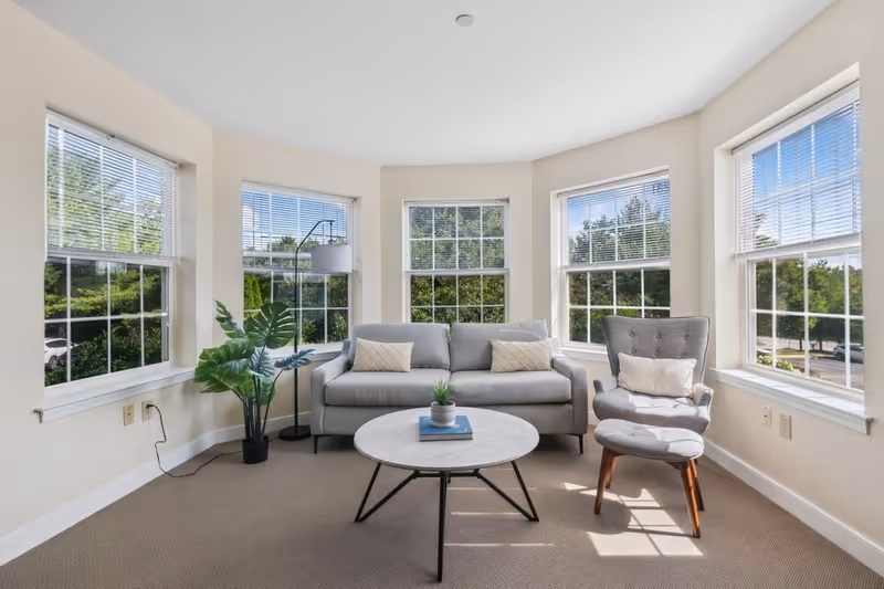A bright living room with large windows on three walls letting in natural light. The room features a gray sofa with two beige pillows, a gray armchair with a matching footrest and a beige pillow, a round coffee table with a small potted plant and a book, a tall floor lamp, and a large green potted plant in the corner.