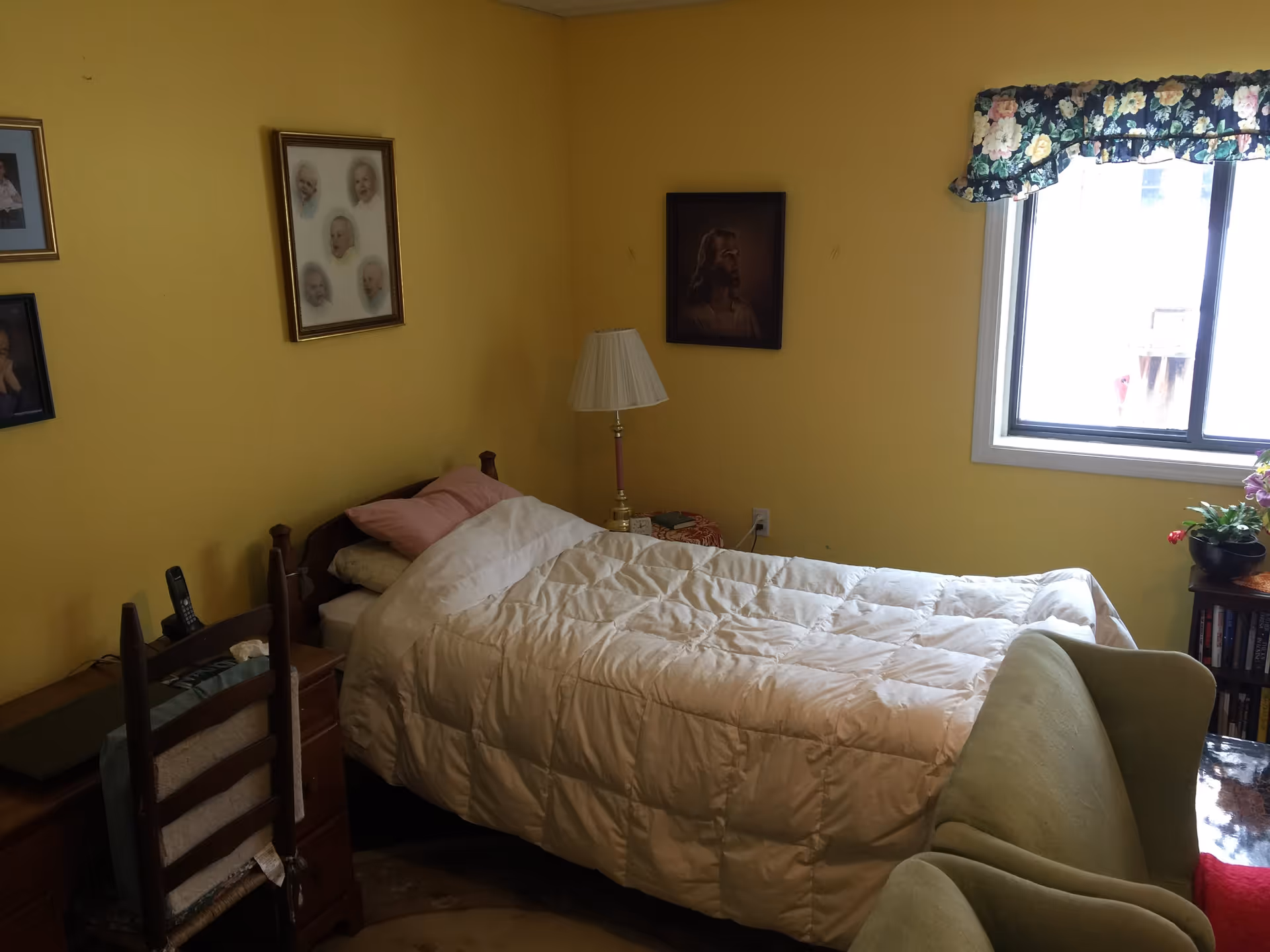 A small bedroom with yellow walls featuring a single bed with a white comforter and pink pillow. There is a wooden desk with a chair, a lamp on a small table, framed pictures on the walls, a window with floral curtains, and a green armchair partially visible.