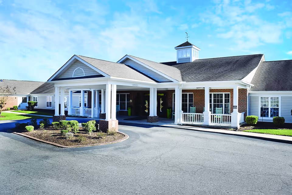 Exterior front view of a single-story senior living facility building with a covered entrance supported by white columns, brick and white siding walls, and a small landscaped area with shrubs in front. The sky is clear and blue.