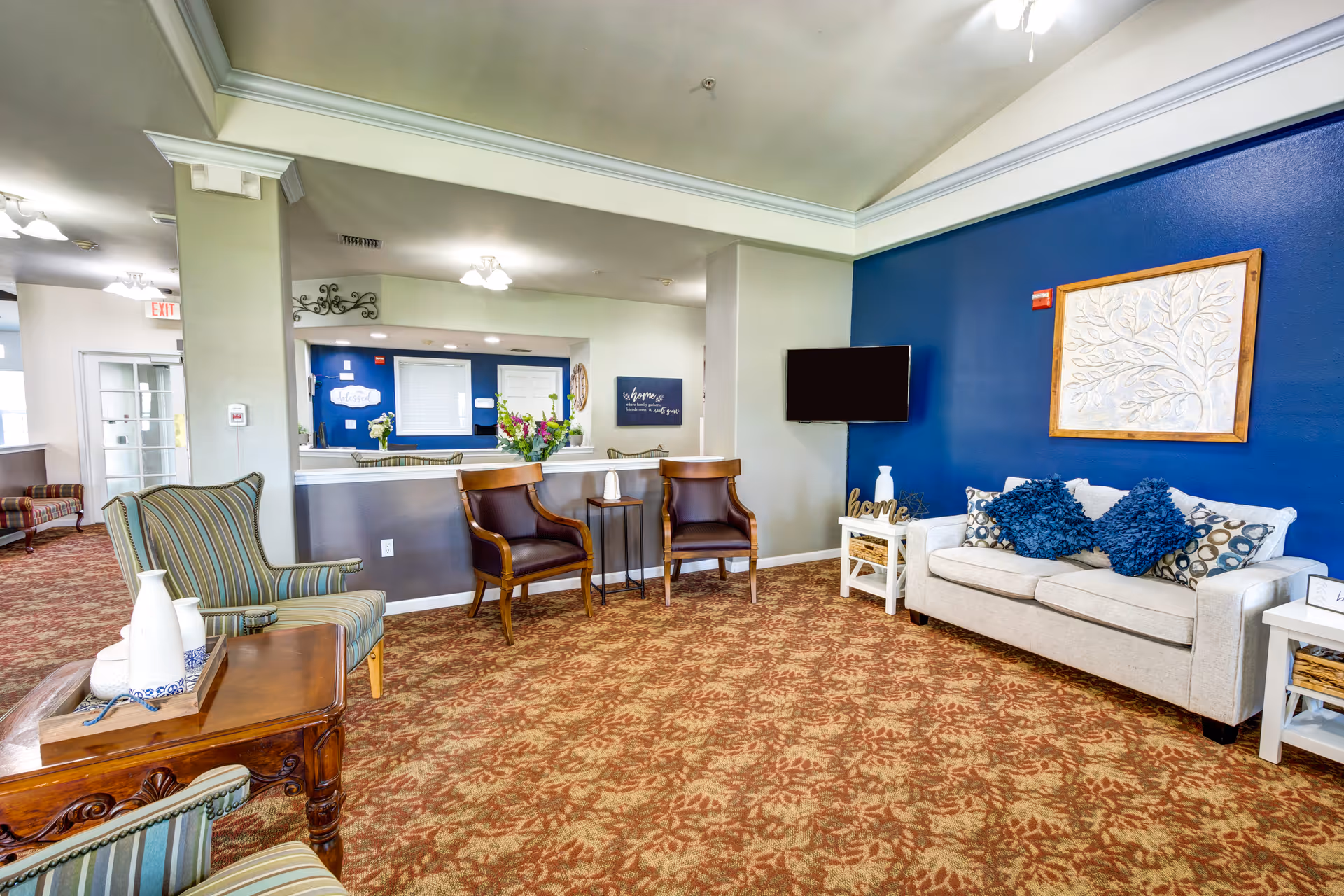A cozy living room area in an assisted living facility featuring a beige sofa with blue and patterned cushions against a blue accent wall with a framed artwork. Two wooden chairs with leather seats are positioned near a small round table. A striped armchair and a wooden side table with decorative vases are also visible. The room has patterned carpet flooring and a wall-mounted TV.