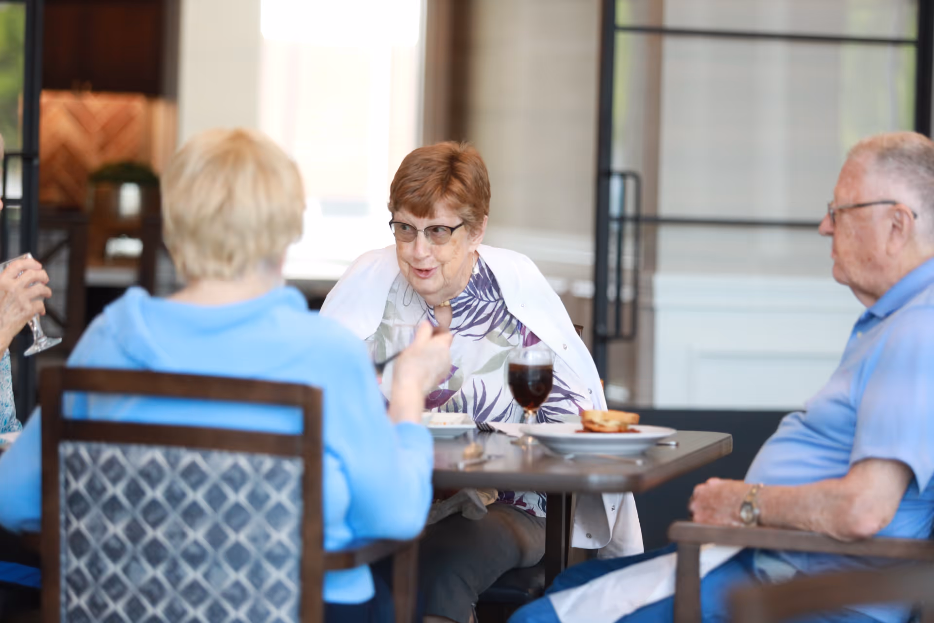 Three elderly people sitting around a dining table engaged in conversation. One woman with short red hair and glasses is facing the camera, while the other two people, a woman in a blue top and a man in a light blue shirt, have their backs or sides to the camera. There are plates and drinks on the table in a well-lit indoor setting.