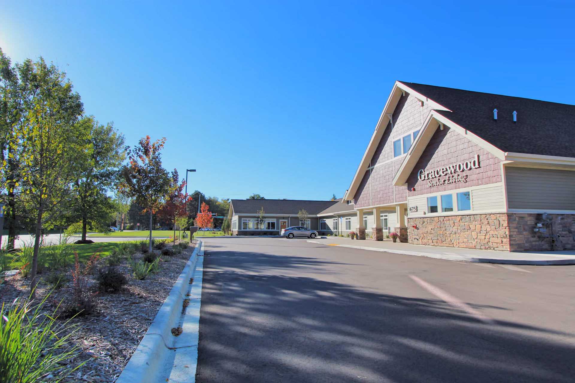 Exterior view of Gracewood Advanced Assisted Living and Memory Care building in Lino Lakes on a clear sunny day, showing the parking lot, landscaped area with trees and shrubs, and the building's stone and siding facade with a peaked roof.