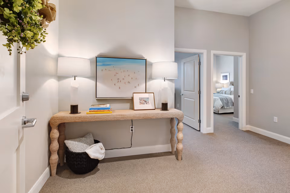 A hallway area with a textured wooden console table against a light gray wall. On the table are two modern table lamps with white shades, a framed photo, and a few stacked books. Above the table hangs a framed artwork depicting a beach scene with small figures. Below the table is a black woven basket containing a gray pillow and a white blanket. To the right, two doorways lead to a bedroom with a neatly made bed and a bedside lamp. The floor is carpeted in a neutral tone.