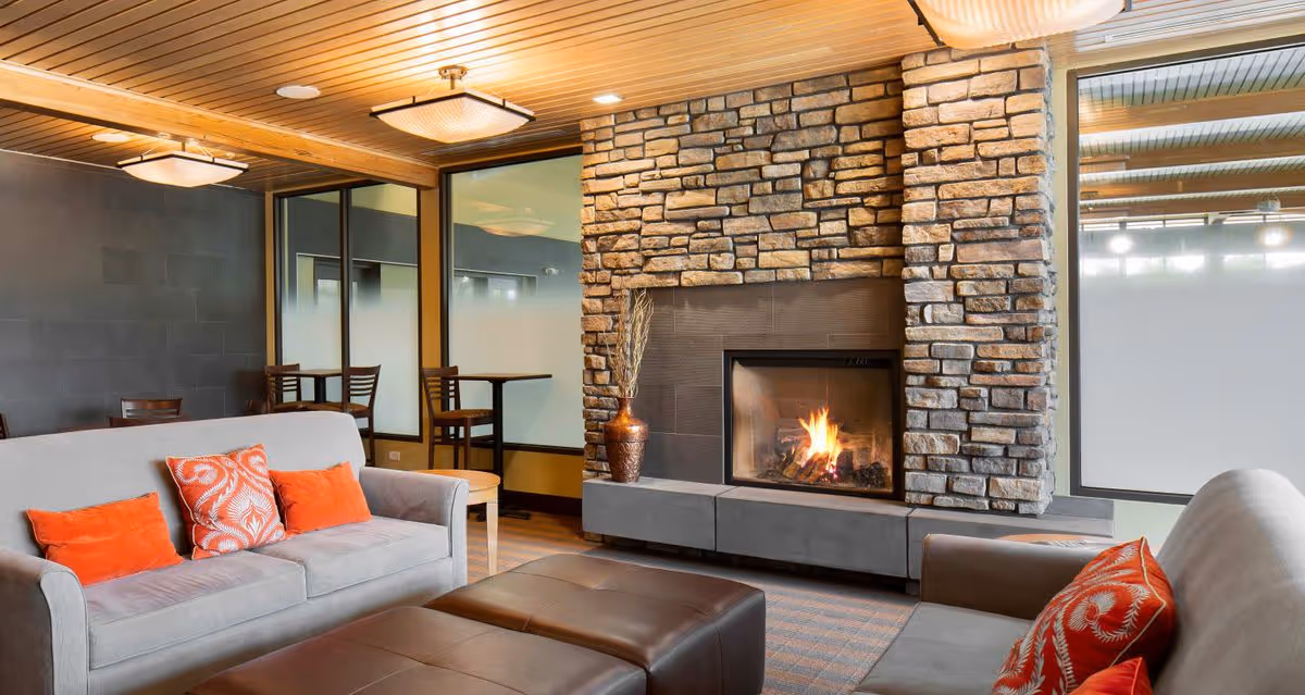 Cozy communal lounge with gray sofas and orange pillows facing a stone fireplace with a burning fire and tables in the background.