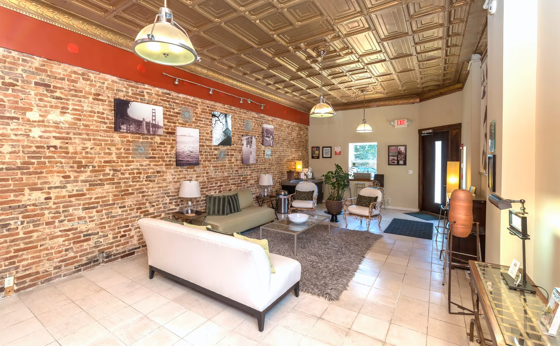 A cozy living room area with a white sofa, a green sofa, two armchairs, a glass coffee table on a shaggy rug, and a brick accent wall decorated with black and white photos. The ceiling has an ornate gold pattern with hanging pendant lights. There is a door and a window at the far end, with some plants and framed pictures on the walls.