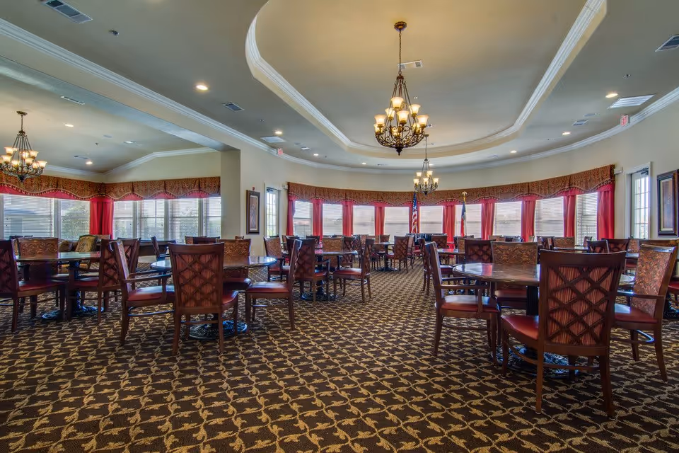 A spacious dining room with multiple round tables and wooden chairs with cushioned seats. The room features patterned carpet flooring, large windows with red curtains and valances, and two ornate chandeliers hanging from a tray ceiling. The walls are light-colored, and there are framed pictures and an American flag near the windows.
