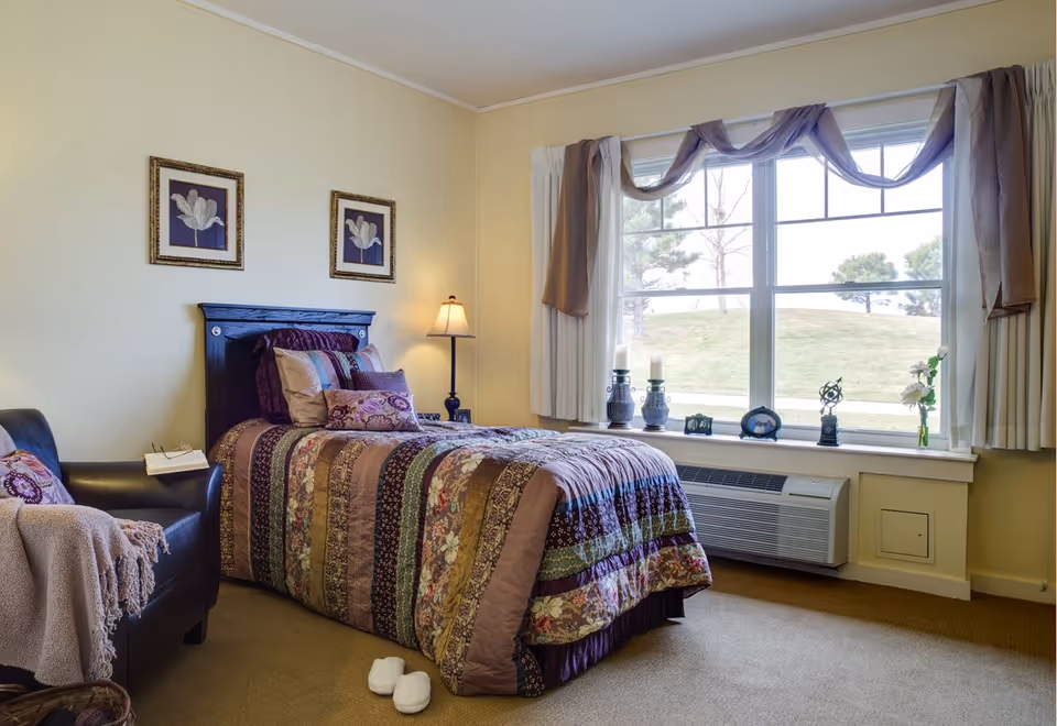 A cozy bedroom with a single bed covered in a colorful patchwork quilt. There are multiple pillows on the bed. Next to the bed is a black armchair with a blanket and a book resting on it. A floor lamp stands beside the bed. The room has a large window with curtains and a windowsill decorated with candles, small framed pictures, and a flower vase. Outside the window, a grassy area with trees is visible.