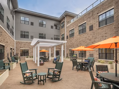 Outdoor courtyard area of a senior living facility with multiple green rocking chairs and tables, some shaded by orange umbrellas. The courtyard is surrounded by a multi-story brick and gray building with many windows. A white pergola structure is centrally located in the courtyard.