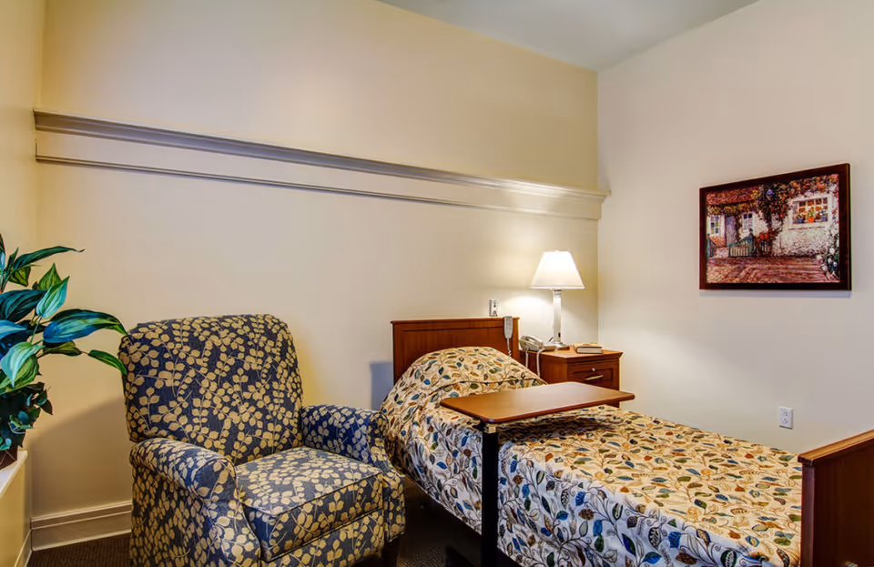 A small bedroom in a senior living facility with a single bed covered in a patterned bedspread, a wooden nightstand with a lamp and telephone, a patterned armchair, a potted plant, and a framed painting on the wall.