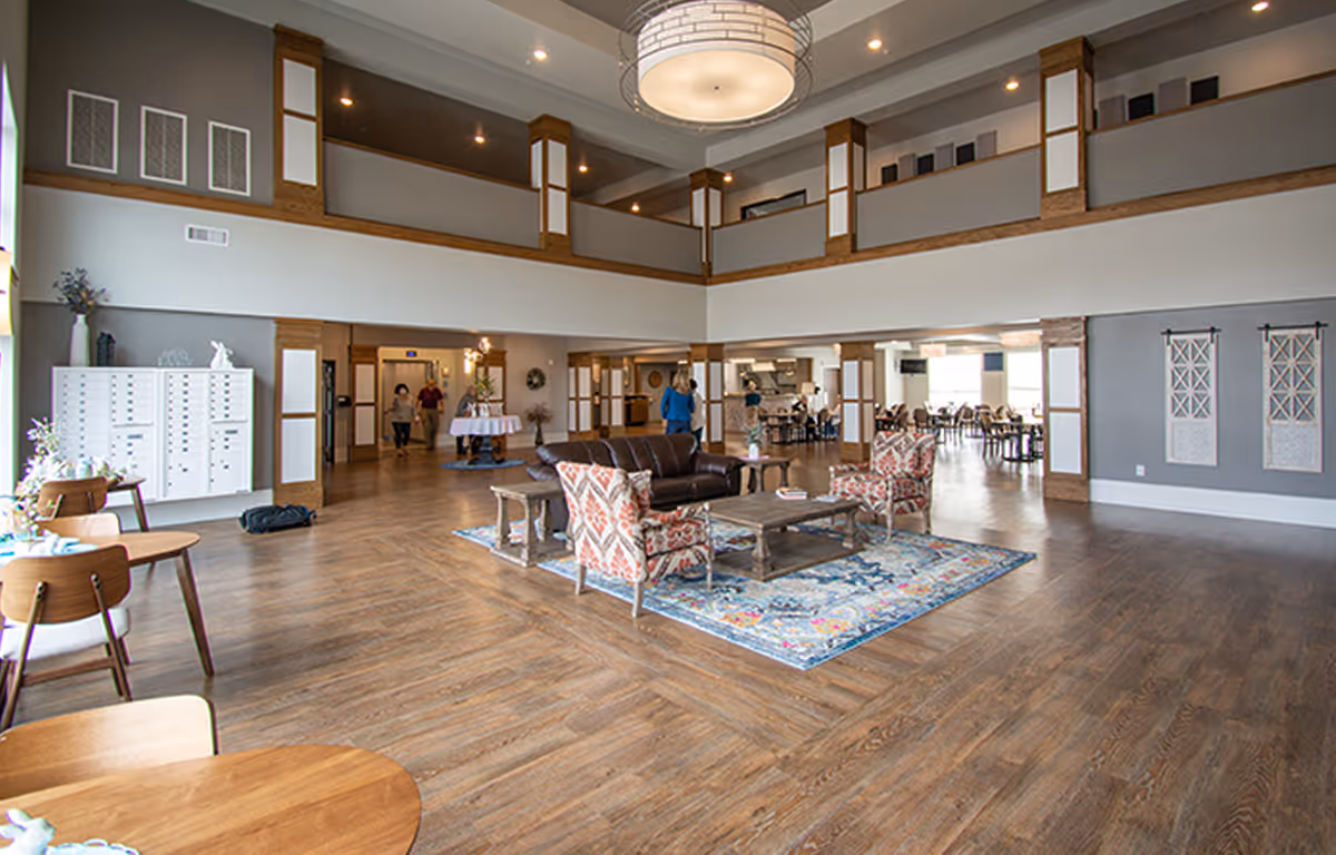 Spacious senior living facility common area with wood flooring, a seating area featuring a leather sofa and patterned armchairs on a colorful rug, round wooden tables with chairs, and a large modern chandelier. The room has high ceilings with a second-floor balcony overlooking the space, and several people are visible in the background near the dining area.