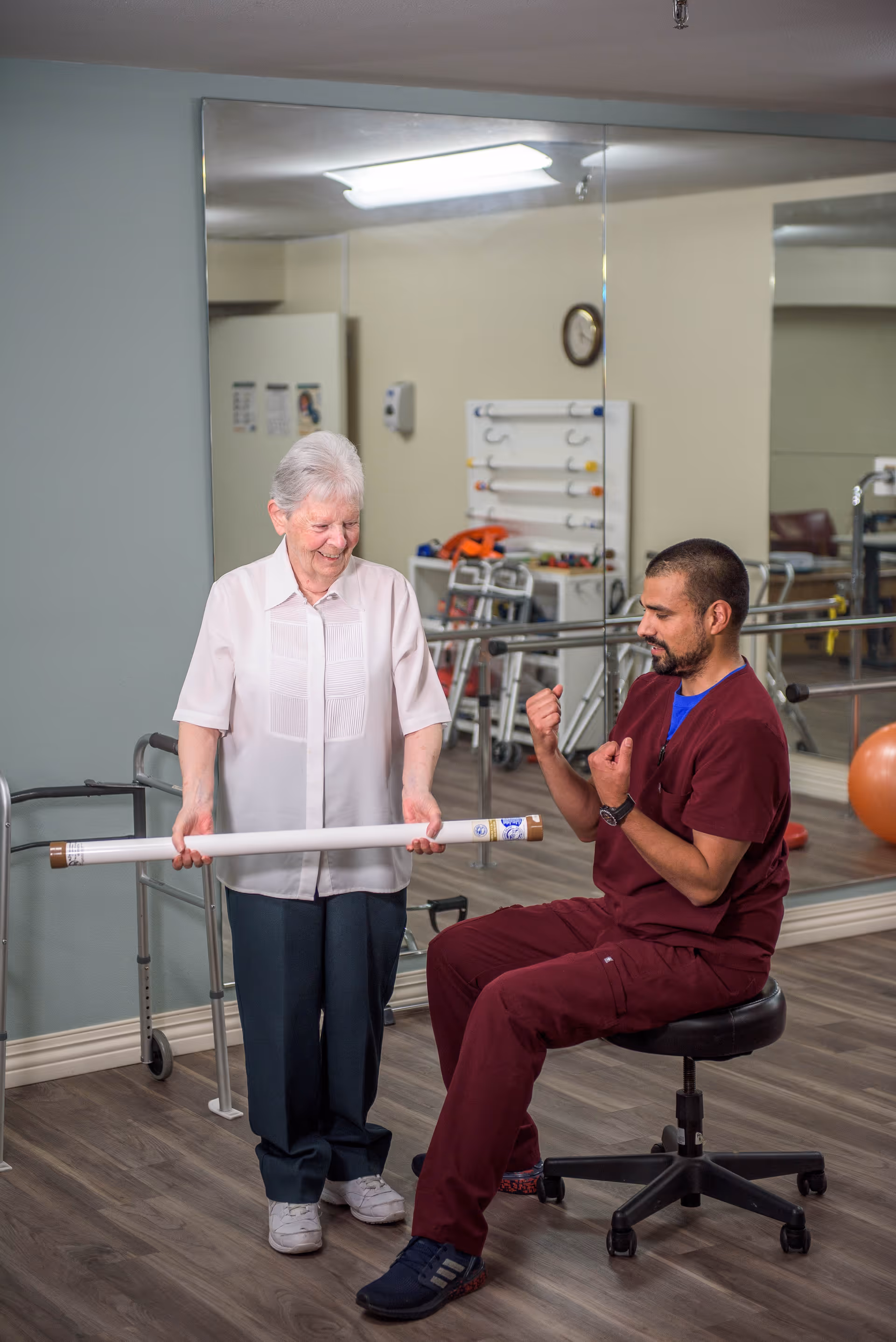 An elderly woman holding a long white tube is standing next to a seated male healthcare worker in maroon scrubs who is making a fist with one hand in a physical therapy or rehabilitation room with mirrors and exercise equipment in the background.