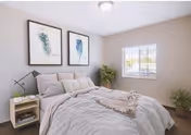 A bright and cozy bedroom with a neatly made bed featuring light-colored bedding and pillows. Two framed botanical prints hang above the bed. There is a small wooden nightstand with a modern lamp and some books on the left side of the bed. A window with white blinds allows natural light to fill the room. Two green plants are placed in the corners, adding a touch of nature to the space.