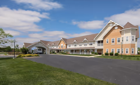 Front exterior of a two-story senior living facility with a covered entrance, driveway, and landscaped lawn under a blue sky.