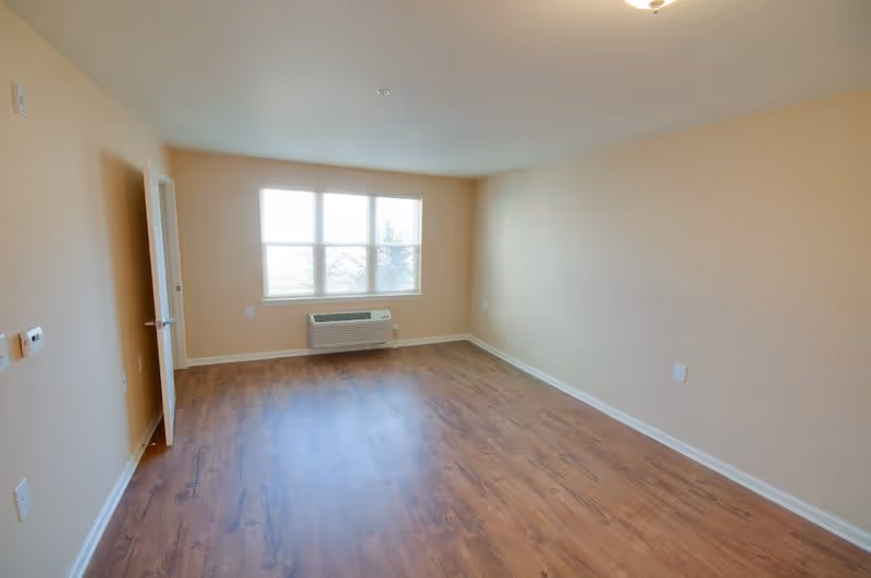 Empty room with beige walls, a large window letting in natural light, a wall-mounted air conditioning unit below the window, wooden flooring, and an open door on the left side.