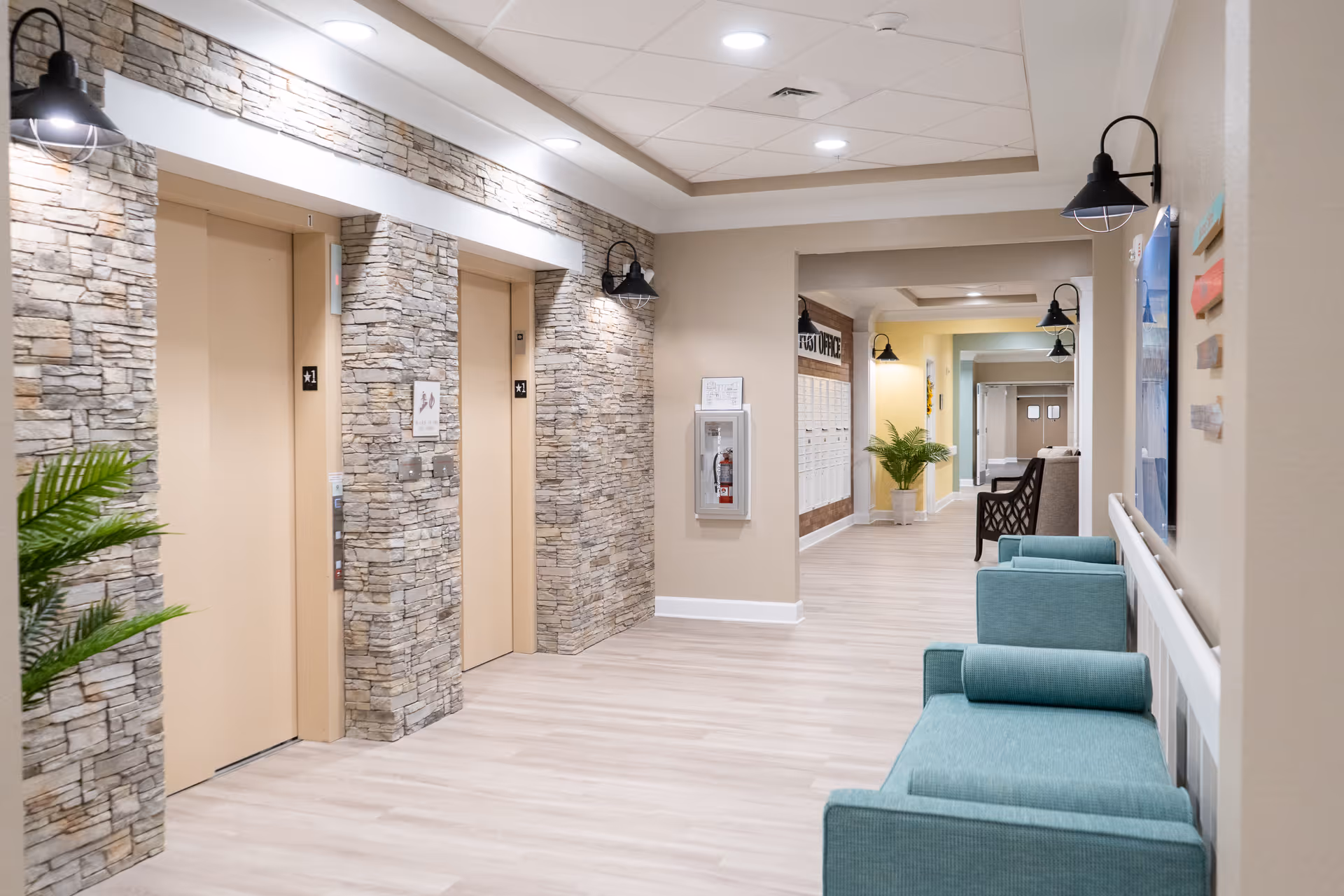 A bright and clean hallway in a senior living facility featuring two beige elevators set in a stone wall, teal upholstered chairs along the right wall, black wall-mounted lamps, a fire extinguisher in a glass case, and a mail area labeled 'POST OFFICE' further down the corridor with potted plants and additional seating.