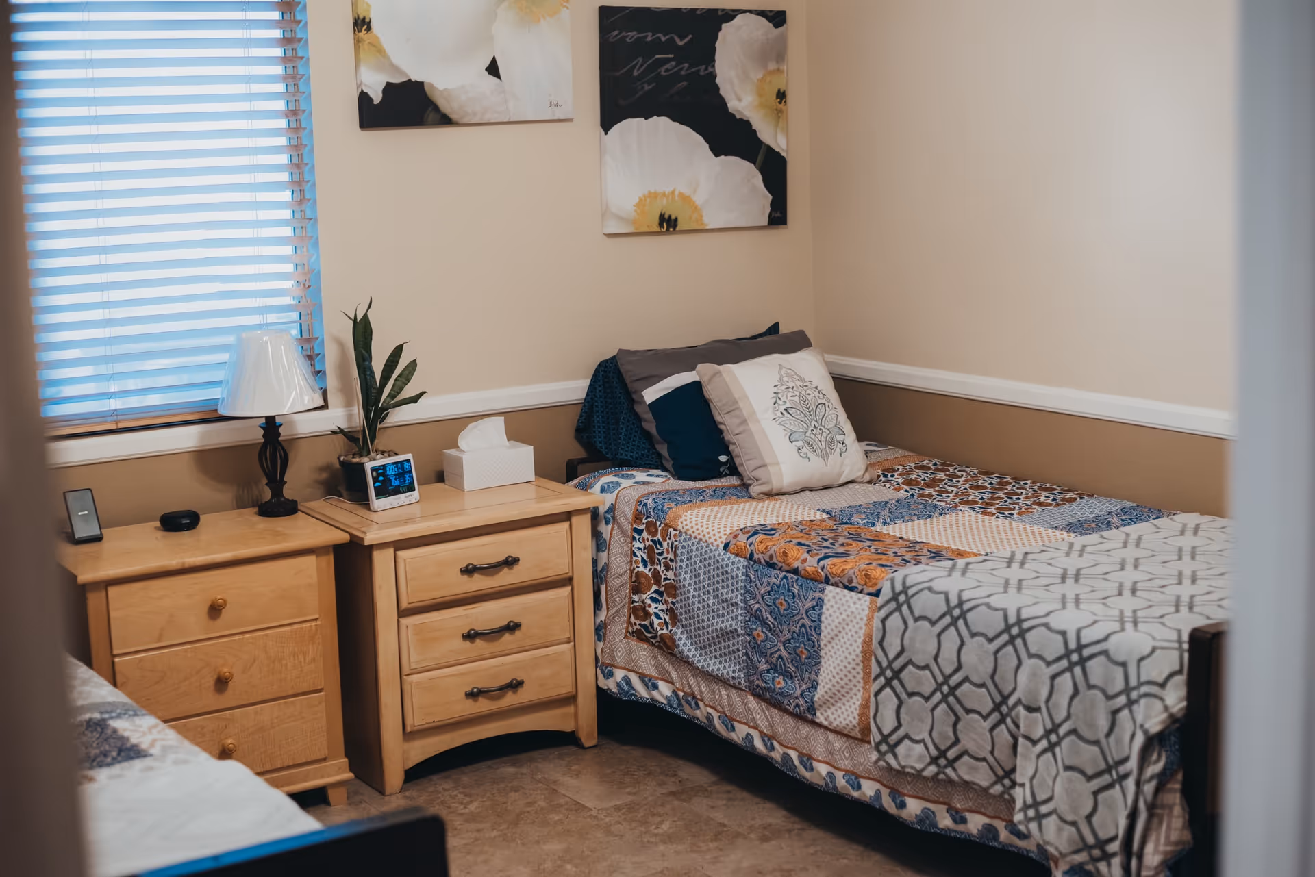 A cozy bedroom in an assisted living facility with a single bed covered in a colorful patchwork quilt and decorative pillows. Next to the bed are two wooden nightstands, one with a lamp, a plant, a tissue box, and a digital clock. The room has beige walls with white trim and two floral paintings hanging above the bed. A window with closed blinds is visible on the left side.