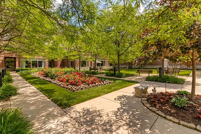 Sunlit landscaped courtyard with paved walkways, flowerbeds, trees, benches, and outdoor seating in front of a brick building.