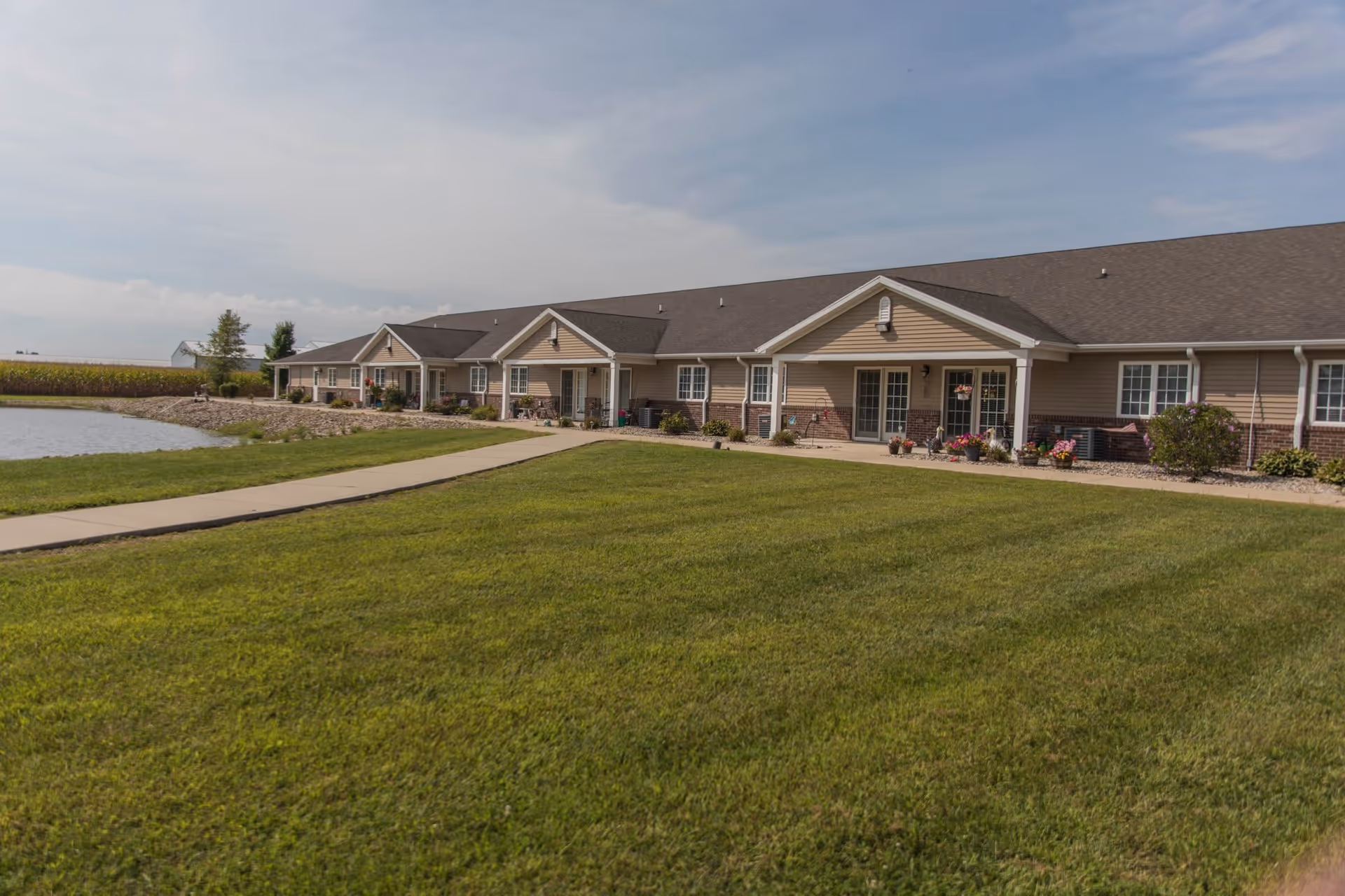 Exterior view of a single-story senior living facility building with beige siding and a dark roof, surrounded by a well-maintained green lawn and a paved walkway leading to the entrance. There is a small pond on the left side and a partly cloudy sky above.