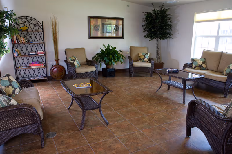 A bright and spacious living room with brown wicker furniture including two armchairs, two sofas, and two glass-top coffee tables. The room has tiled flooring, a large window letting in natural light, a bookshelf with books and decor, potted plants, and a framed artwork on the wall.