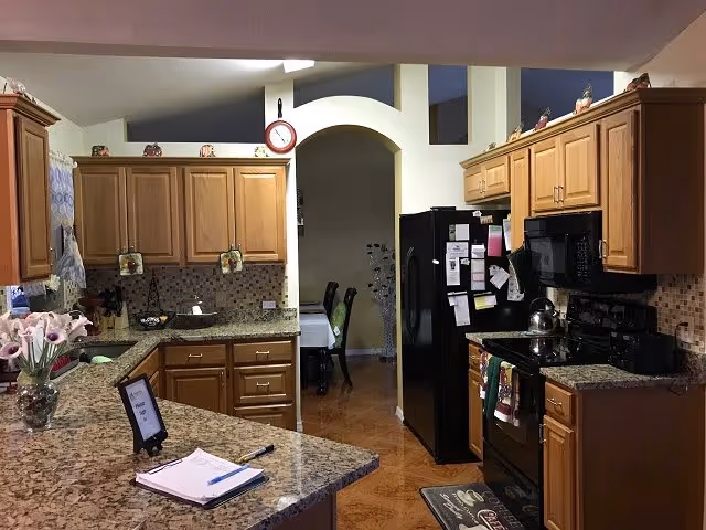Interior view of a kitchen with wooden cabinets, granite countertops, a black refrigerator, stove, and microwave. There is a dining area visible through an arched doorway in the background. Various kitchen items and decorations are placed on the counters and cabinets.