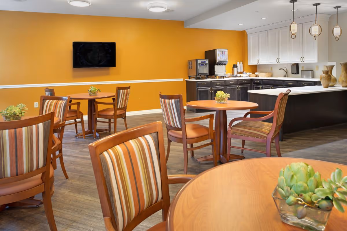 Dining room with round wooden tables and striped chairs, a kitchenette with white cabinets, and a TV mounted on an orange wall.