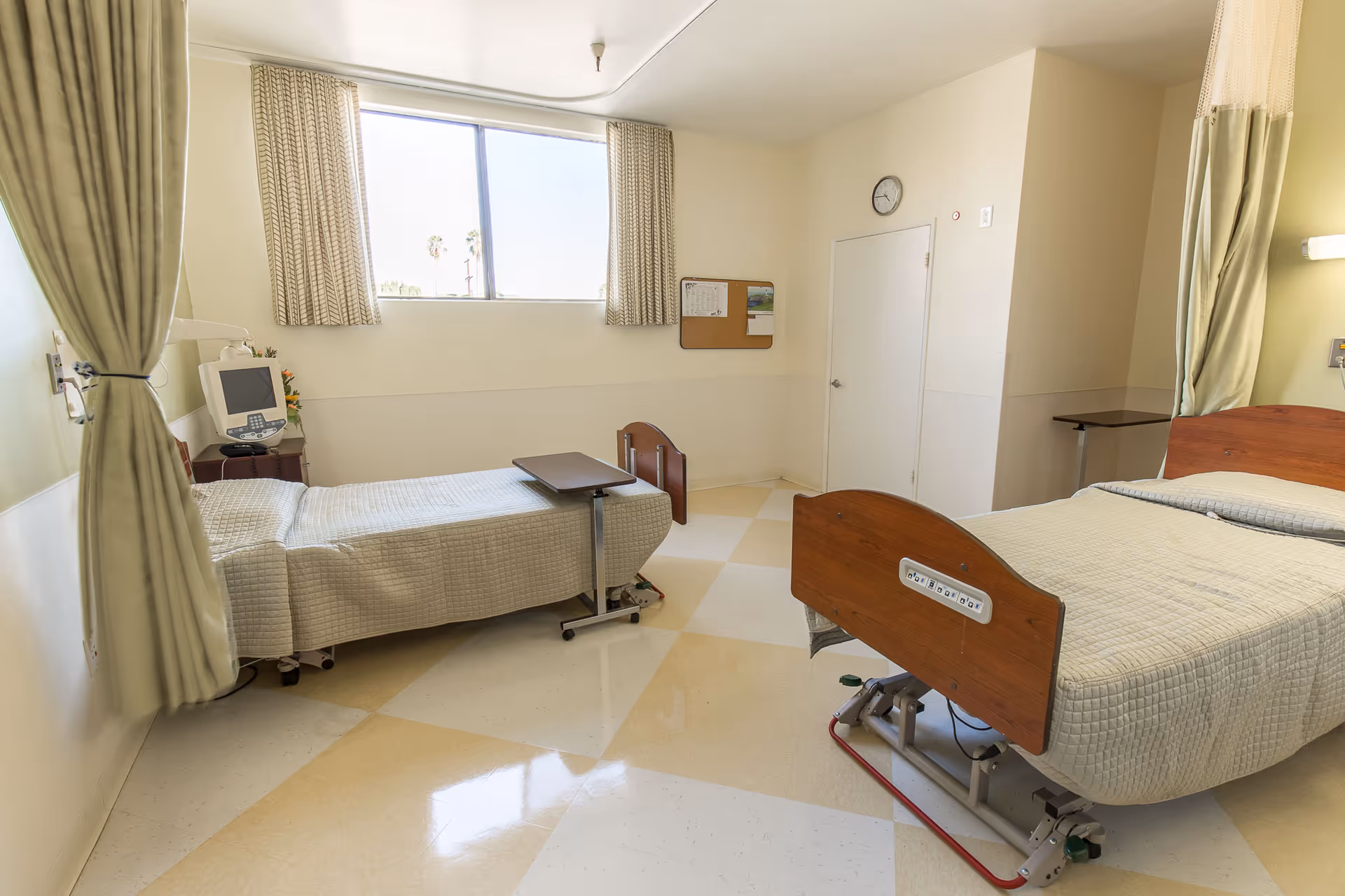 A bright hospital room with two adjustable beds covered in beige bedspreads, a small overbed table, a medical monitor on a stand, a window with curtains, a clock on the wall, and a bulletin board.
