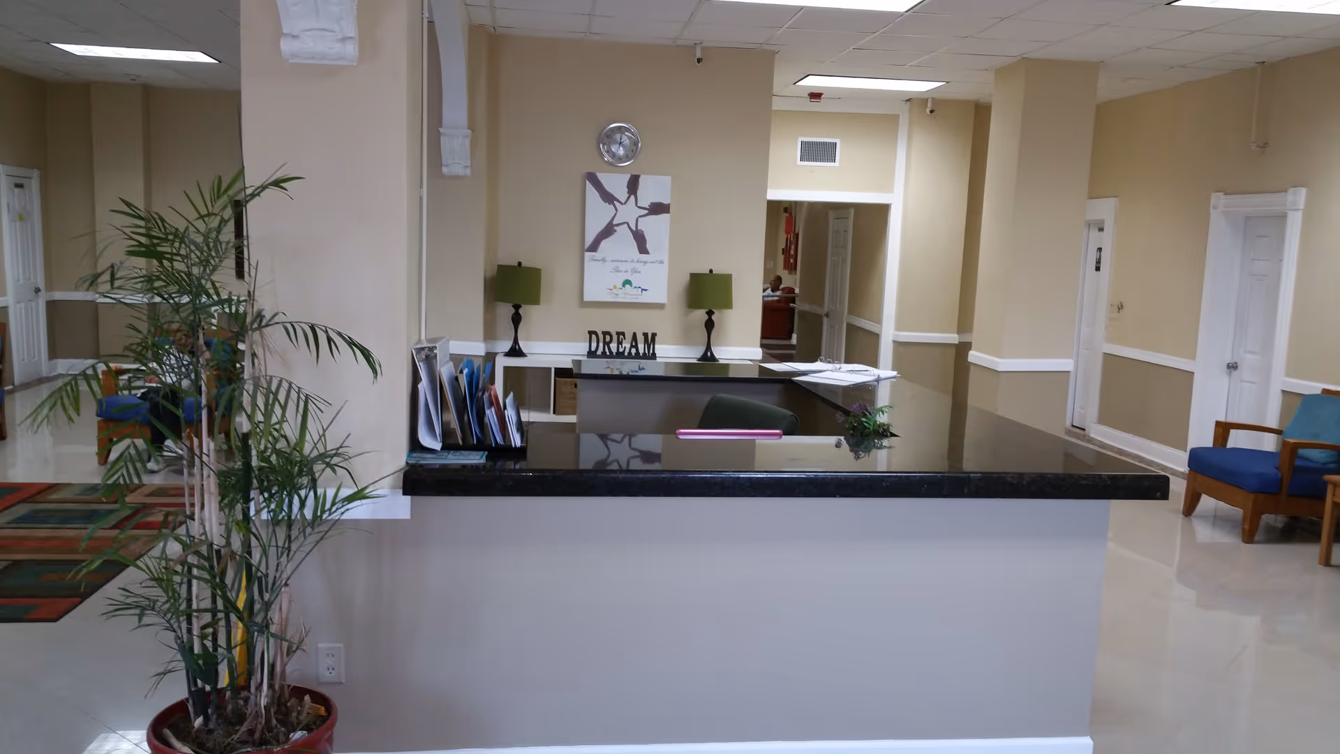 Reception desk in a bright facility lobby with potted plants, seating, lamps, and a 'DREAM' sign on the back wall.
