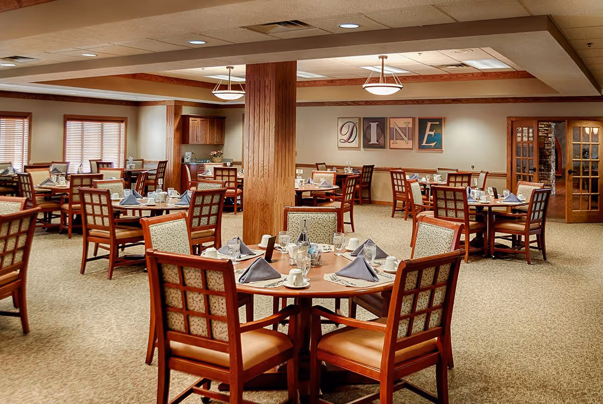 A spacious dining room in a senior assisted living facility with multiple round and square wooden tables set with plates, cups, glasses, and folded napkins. The room has beige carpet, wooden trim, and soft lighting from ceiling fixtures. On the far wall, there are four framed letters spelling 'DINE'.