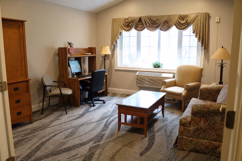 A cozy living room with a patterned carpet, a wooden coffee table with a glass top, a floral upholstered sofa, a beige armchair, a floor lamp, and a wooden desk with a computer and office chair near a window with beige curtains.