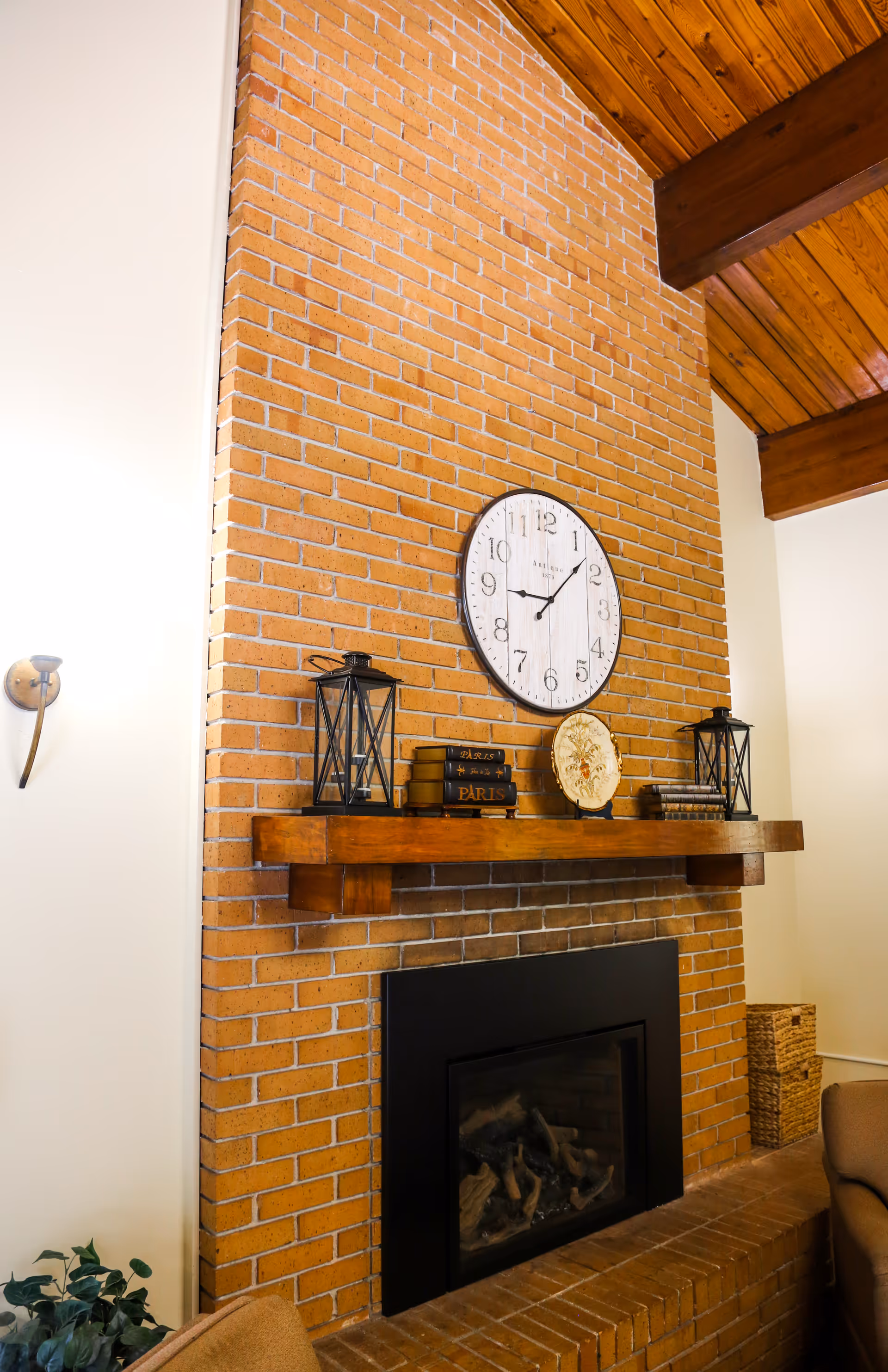 A cozy brick fireplace with a wooden mantel decorated with two black lanterns, a stack of books, and a decorative plate. Above the mantel is a round wall clock with black numbers and hands. The room has a wooden ceiling with exposed beams and a beige wall with a wall sconce light fixture.