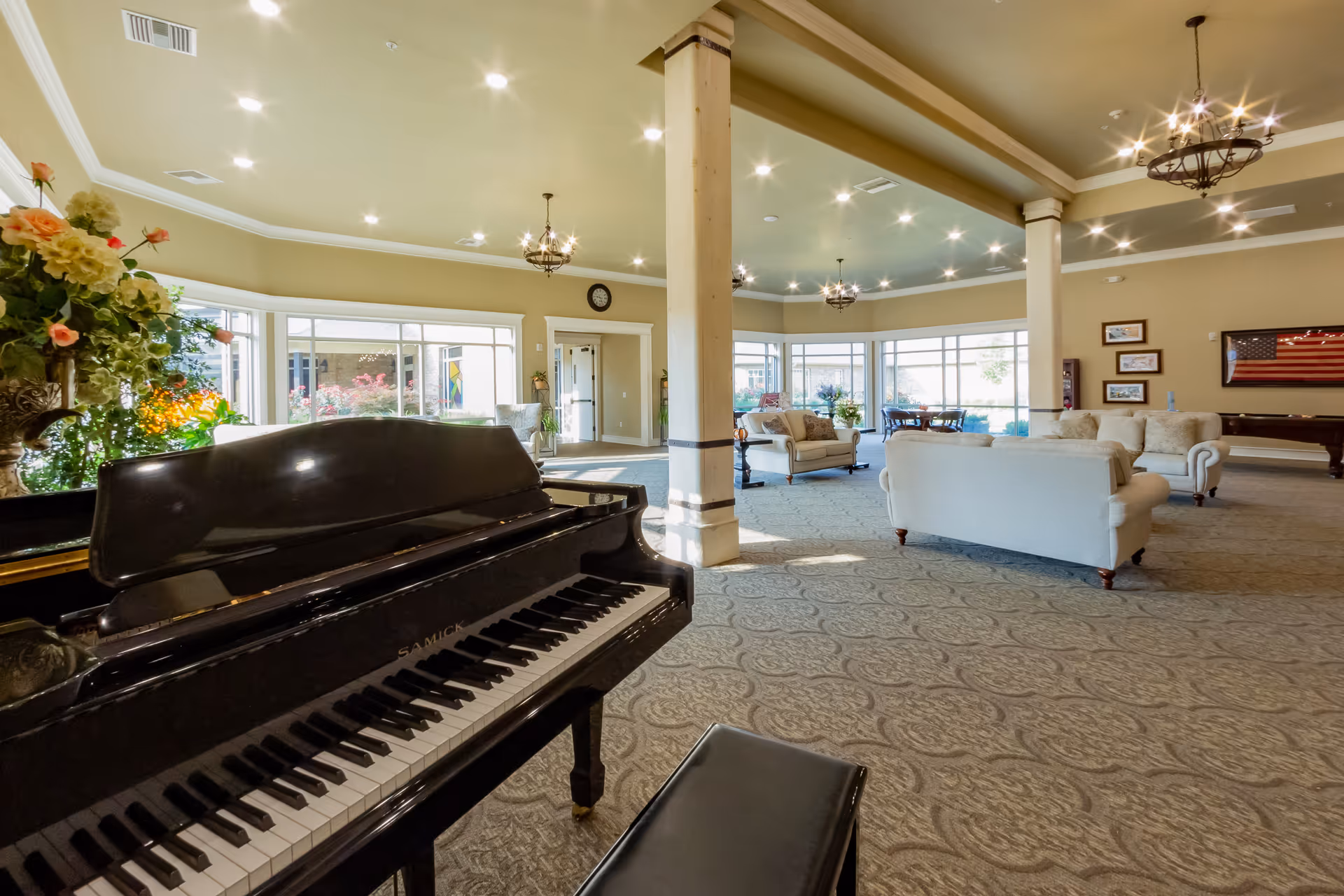 Spacious and well-lit common area with a black grand piano in the foreground, cream-colored sofas, large windows letting in natural light, decorative columns, chandeliers, and framed pictures on the wall.