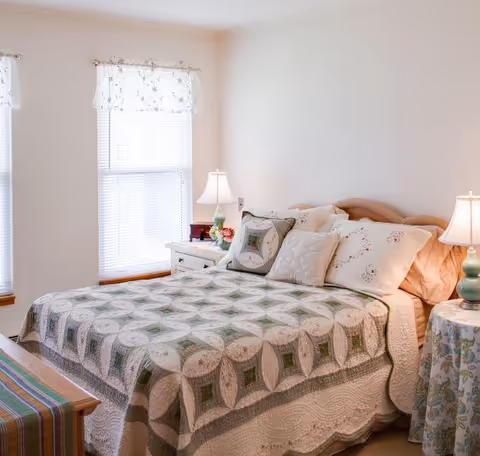 A cozy bedroom with a bed covered in a patterned quilt and multiple pillows. There are two bedside tables with lamps on each side of the bed. The room has two windows with white blinds and floral curtains, and light-colored walls.