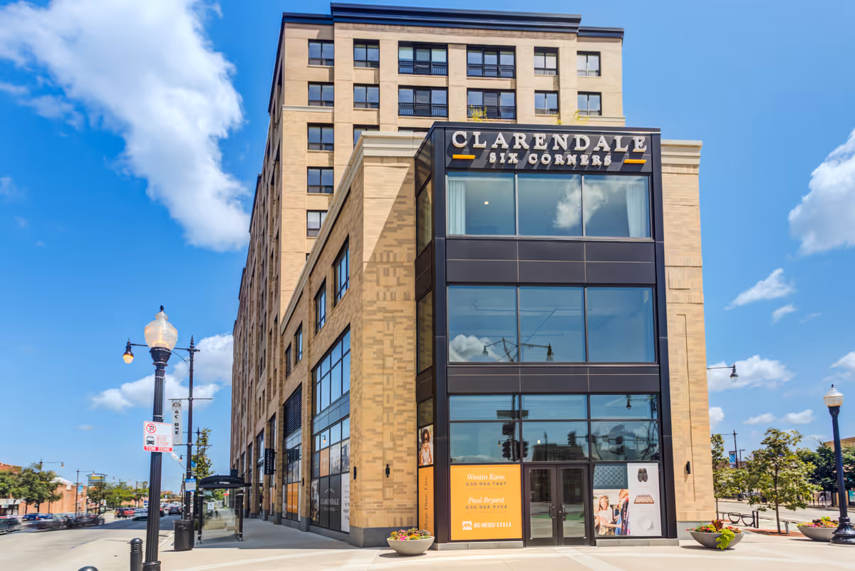 Exterior view of a multi-story building named Clarendale Six Corners on a sunny day with a clear blue sky and some clouds. The building has large windows and a modern design with beige brick and dark accents. There are street lamps, a bus stop, and some trees along the sidewalk.