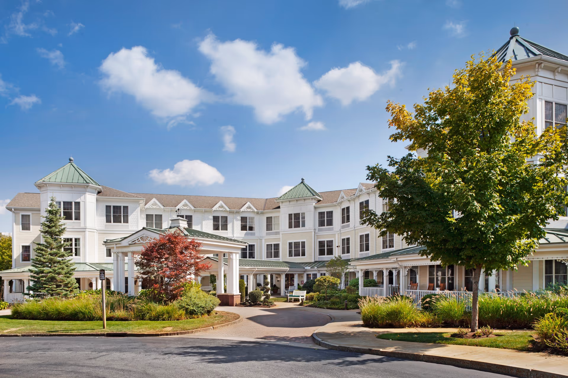 Front exterior of a multi-story white senior living facility with a covered entrance, landscaped grounds, and trees under a blue sky.
