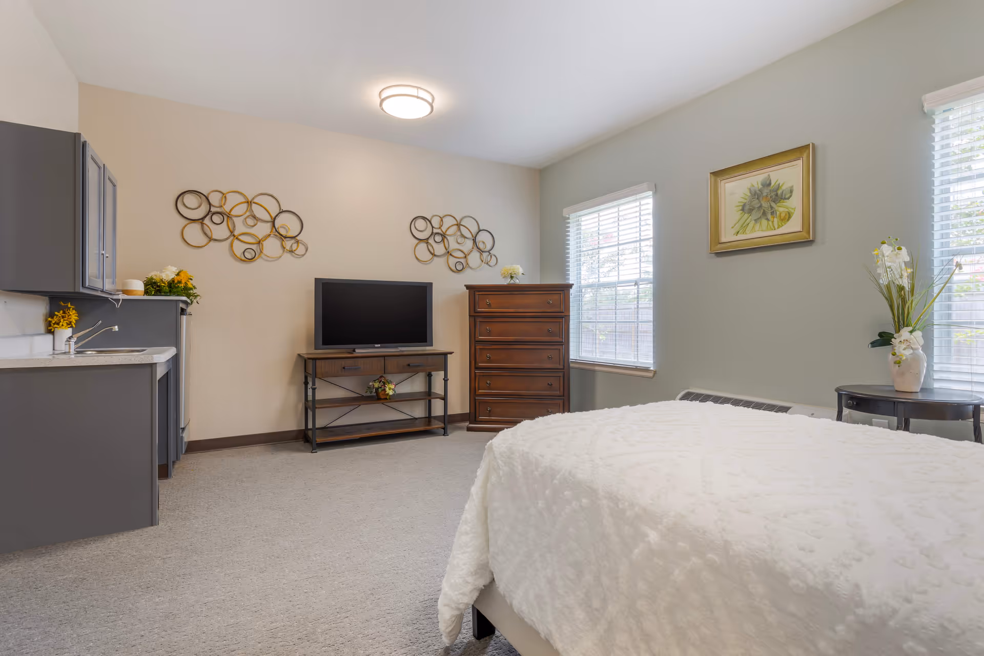 A senior living facility bedroom with a bed covered in a white textured blanket, a wooden dresser, a TV on a wooden stand, two windows with blinds, a small round table with a vase of flowers, and a kitchenette with gray cabinets and a sink.