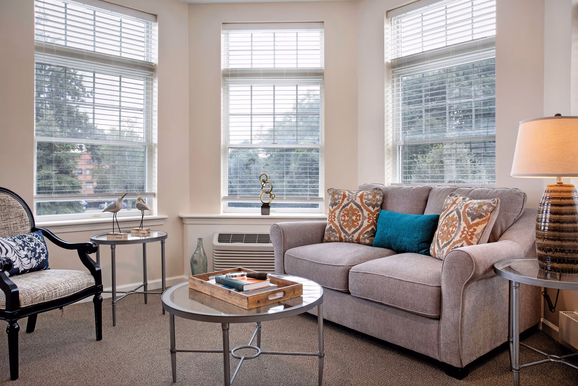A cozy living room with a gray sofa adorned with patterned and teal throw pillows, a glass-top coffee table with a wooden tray holding books, a side table with a lamp, an armchair with a decorative pillow, and three large windows with white blinds letting in natural light.