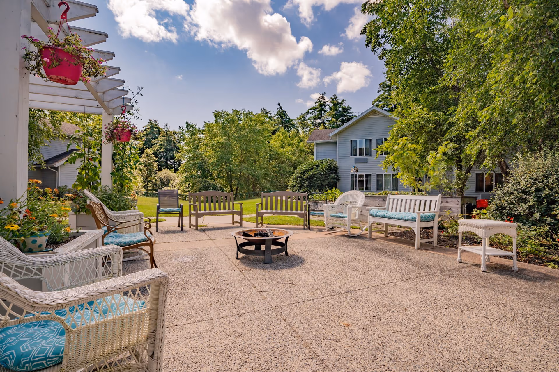 Outdoor patio area with multiple chairs and benches arranged around a central fire pit. The patio is surrounded by greenery including trees and hanging flower pots, with a building visible in the background under a partly cloudy sky.