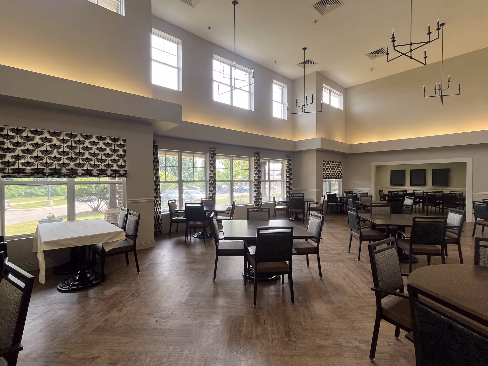 A spacious dining room with multiple tables and chairs arranged neatly. The room features large windows with patterned curtains allowing natural light to fill the space. The ceiling is high with modern chandeliers and recessed lighting along the perimeter. The floor has a wood-like finish.