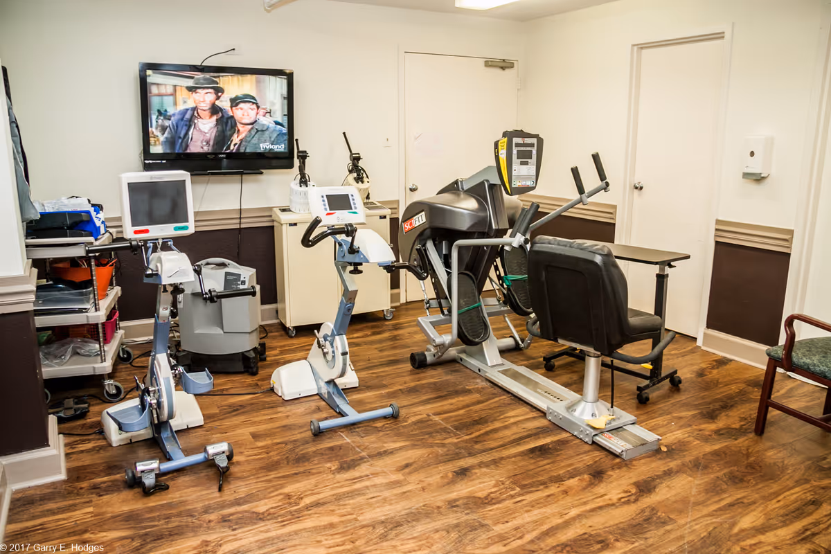 An interior therapy room with exercise bikes, a recumbent machine, chairs, and a wall-mounted TV.