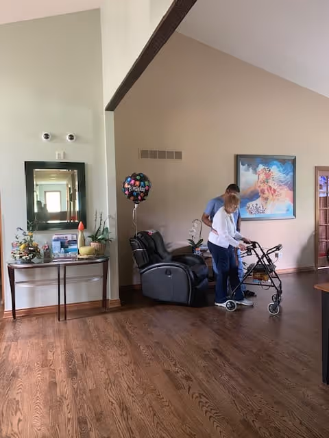 A spacious room with wooden flooring featuring a black recliner chair, a side table with decorative items and plants, and a colorful balloon. Two people are present; one is assisting the other who is using a walker. A large framed artwork hangs on the beige wall in the background.
