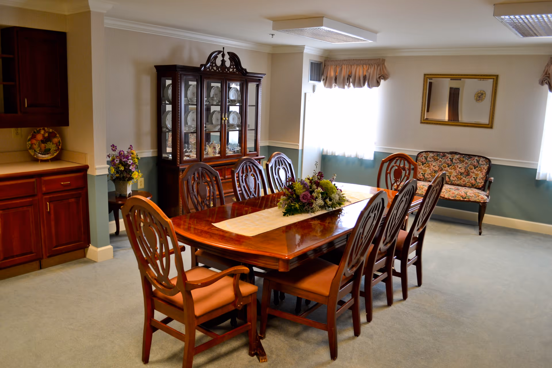 A traditional dining room with a polished wooden dining table surrounded by eight matching chairs with cushioned seats. A floral centerpiece is placed on a table runner in the middle of the table. Behind the table is a glass-fronted china cabinet displaying plates and decorative items. To the right, there is a floral upholstered bench under a wall mirror, and two windows with curtains letting in natural light. On the left side, wooden cabinets and a small table with a flower arrangement are visible.