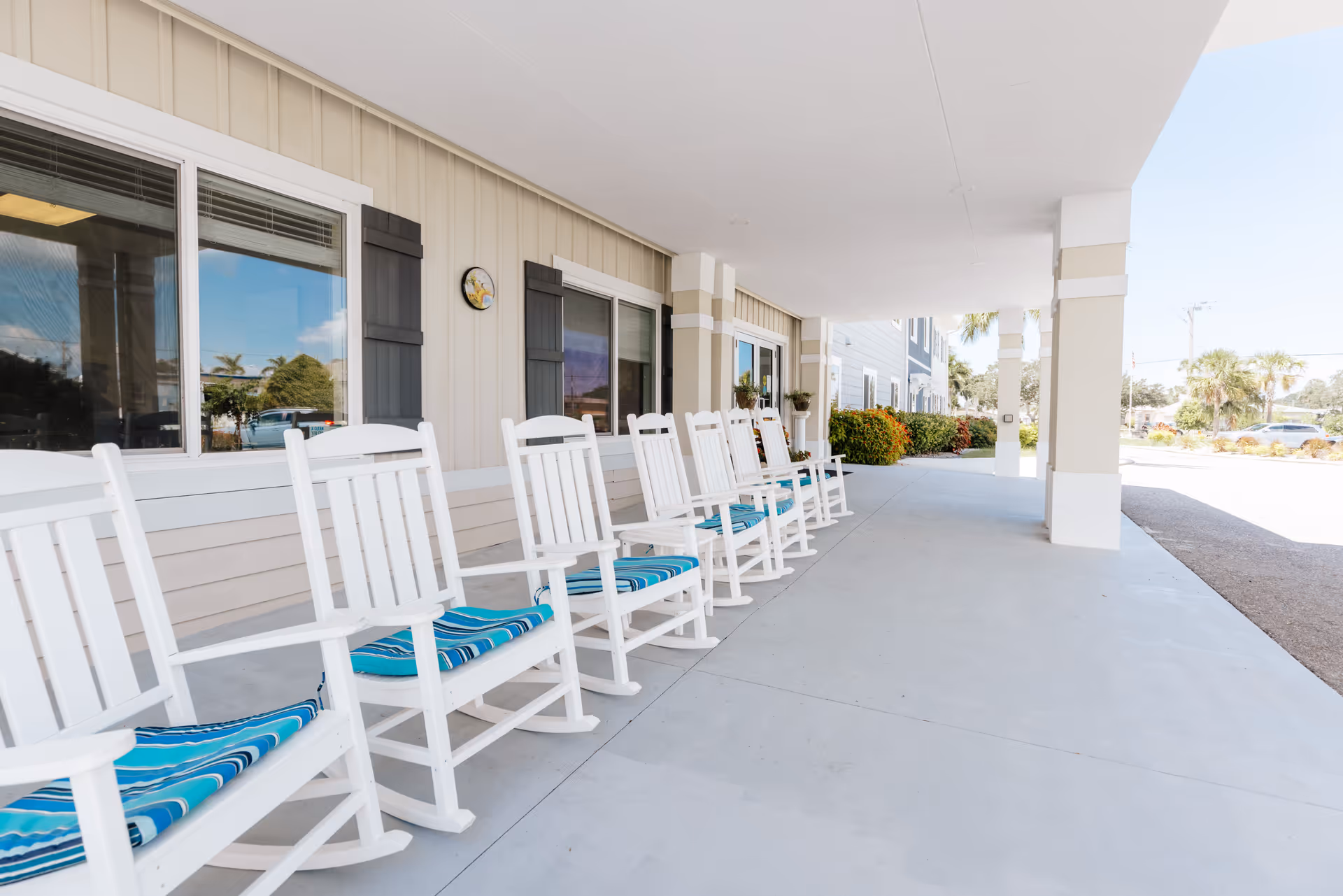 A row of white wooden rocking chairs with blue striped cushions lined up on a covered porch outside a building with beige siding and black window shutters. There are windows and a clock on the wall, and some greenery and palm trees are visible in the background.