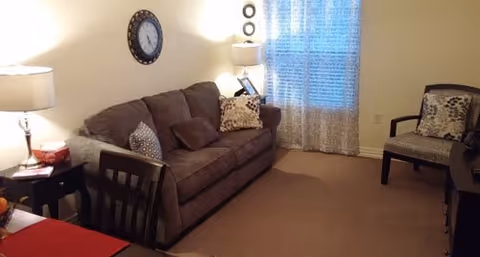 A cozy living room with a brown sofa adorned with patterned cushions, a wooden chair with a cushion, a side table with a lamp, a wall clock, and a window with sheer curtains letting in natural light.