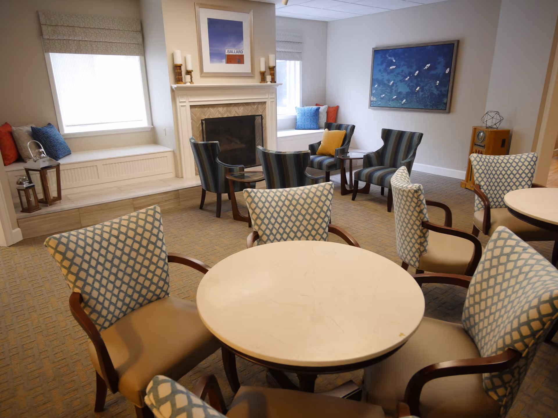 A cozy common area with a round table surrounded by six cushioned chairs with patterned backs. In the background, there are four striped armchairs arranged around a fireplace with candles on the mantel. Two windows with blinds and cushioned window seats with colorful pillows flank the fireplace. A framed picture hangs above the fireplace, and another large framed artwork featuring boats on water is on the adjacent wall. The room has a warm and inviting atmosphere.