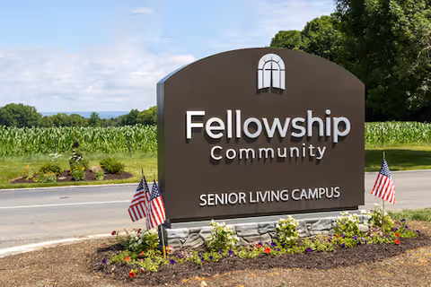 Entrance sign reading "Fellowship Community Senior Living Campus" surrounded by landscaping and small American flags with a road and fields behind it.