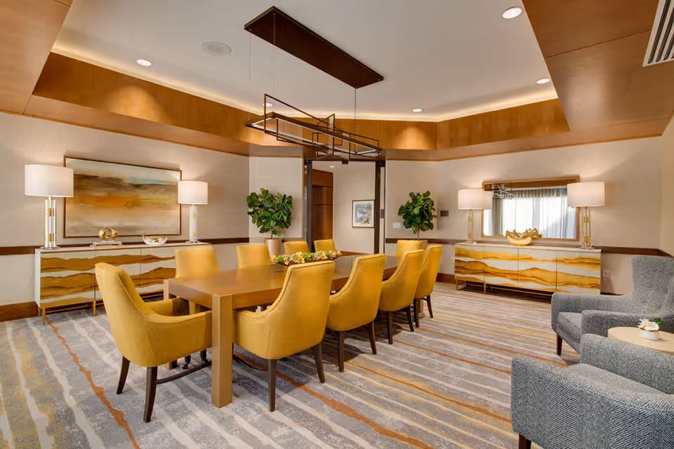 A modern dining room with a large wooden table surrounded by eight mustard yellow upholstered chairs. The room features two matching sideboards with abstract yellow and white patterns, each topped with two lamps and decorative items. Two green plants are placed near the walls, and a large abstract painting hangs above one sideboard. The ceiling has recessed lighting and a contemporary rectangular chandelier above the table. The floor is covered with a patterned carpet in shades of gray, beige, and orange. Two gray upholstered armchairs with a small round table are visible in the foreground.