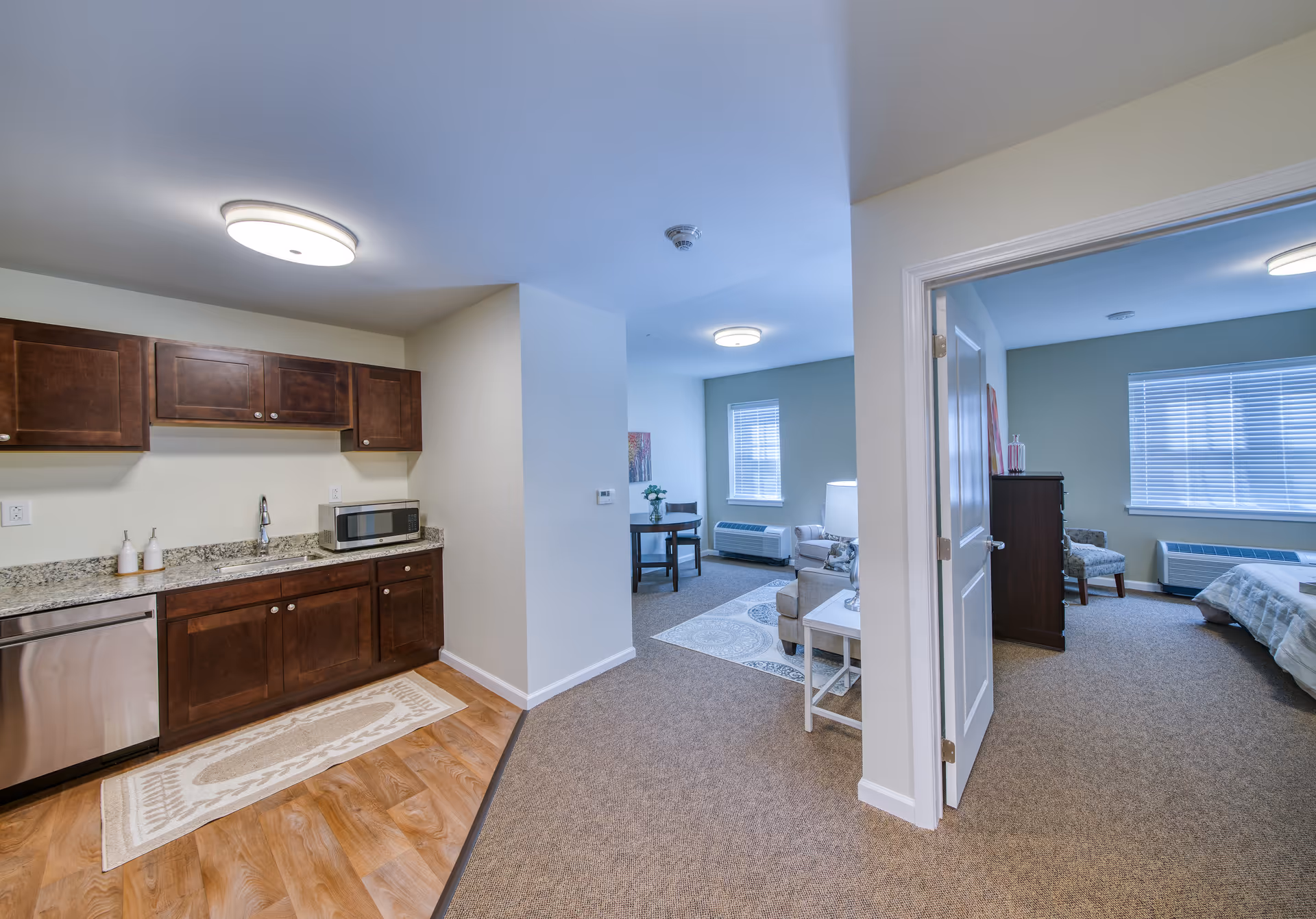Interior view of a senior living apartment showing a small kitchen area with dark wooden cabinets, granite countertop, microwave, and dishwasher on the left. The kitchen opens into a living room with a chair, side table, and a small dining table with two chairs near a window. To the right, there is an open door leading to a bedroom with a bed, dresser, and chair, illuminated by natural light from a window with blinds.